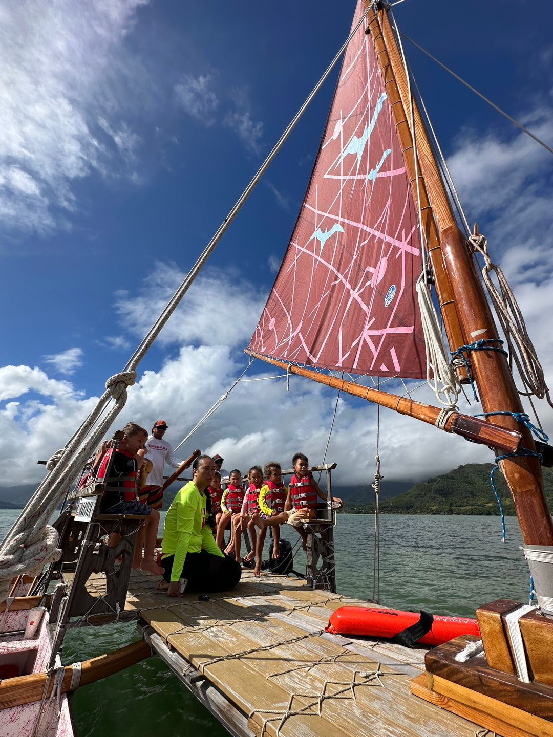 School group on a waa canoe with a red sail on kaihekauila against a blue sky.
