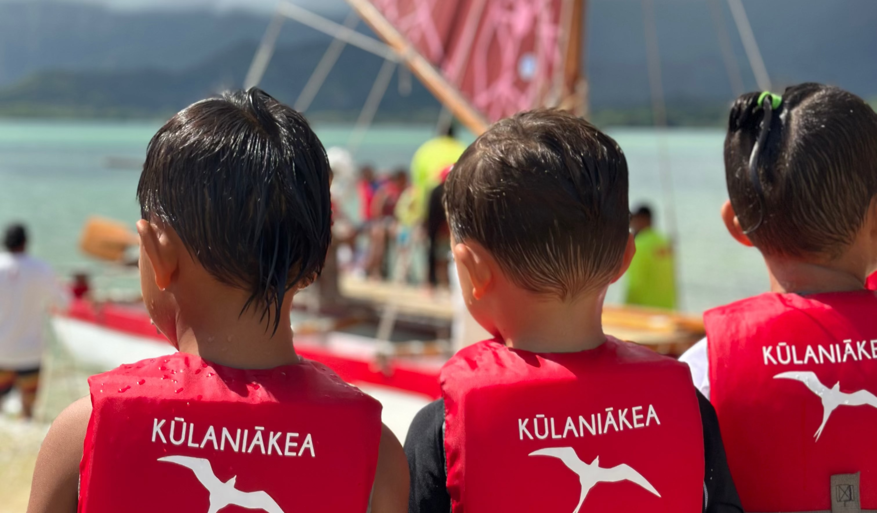 Three children wearing red life vests with 'Kulaniakea'" on a beach with a traditional sailboat in the background.